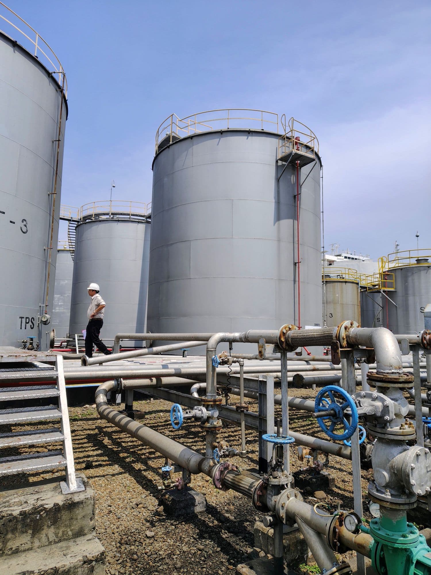 Person in a white hard hat walking between large storage tanks and pipework
